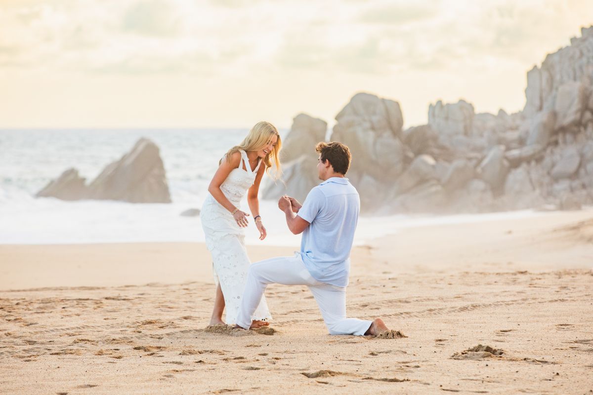 Man proposing on one knee to a woman in a white dress on a sandy beach at sunset.