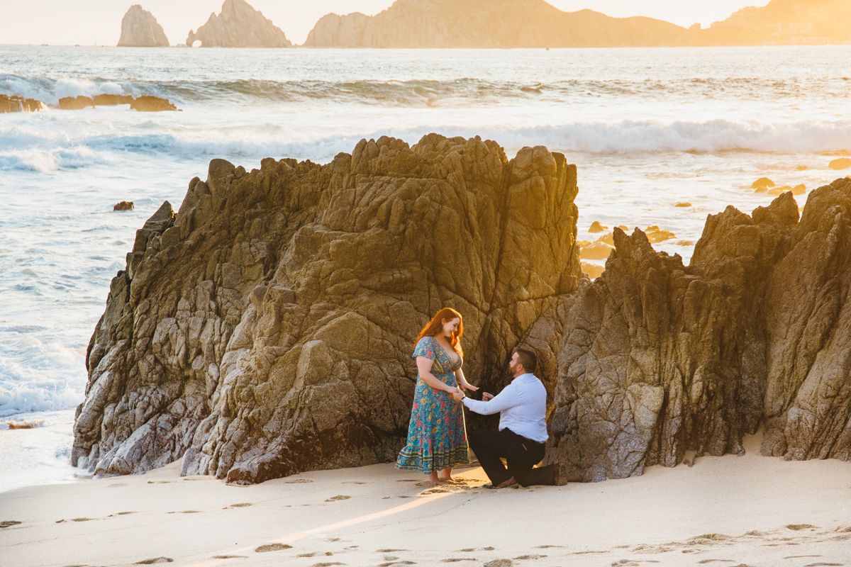 Man proposing to woman on sandy beach near dramatic rock formations with ocean waves at golden sunset.