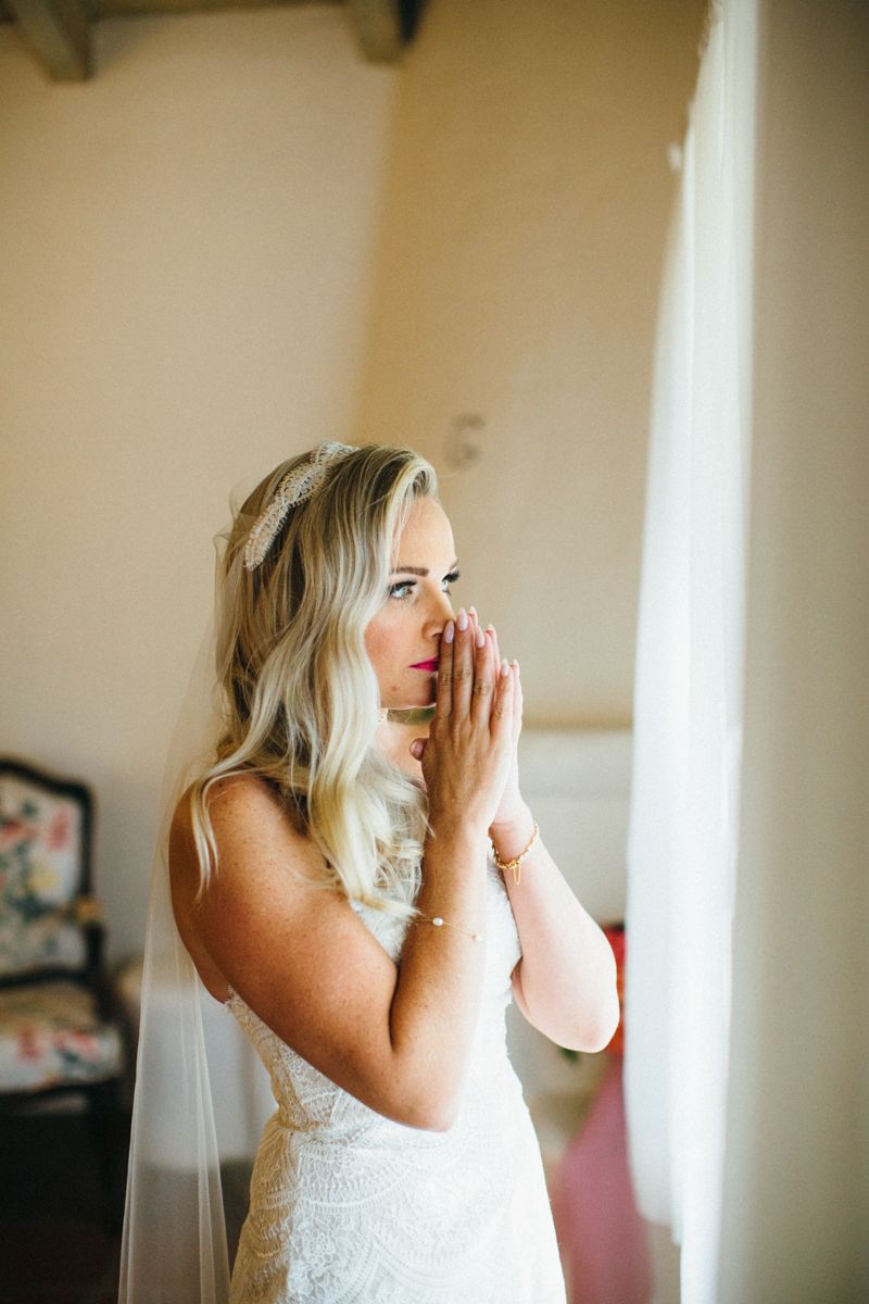 A blonde bride in a lace gown and veil gazes out a window with hands clasped before her wedding.