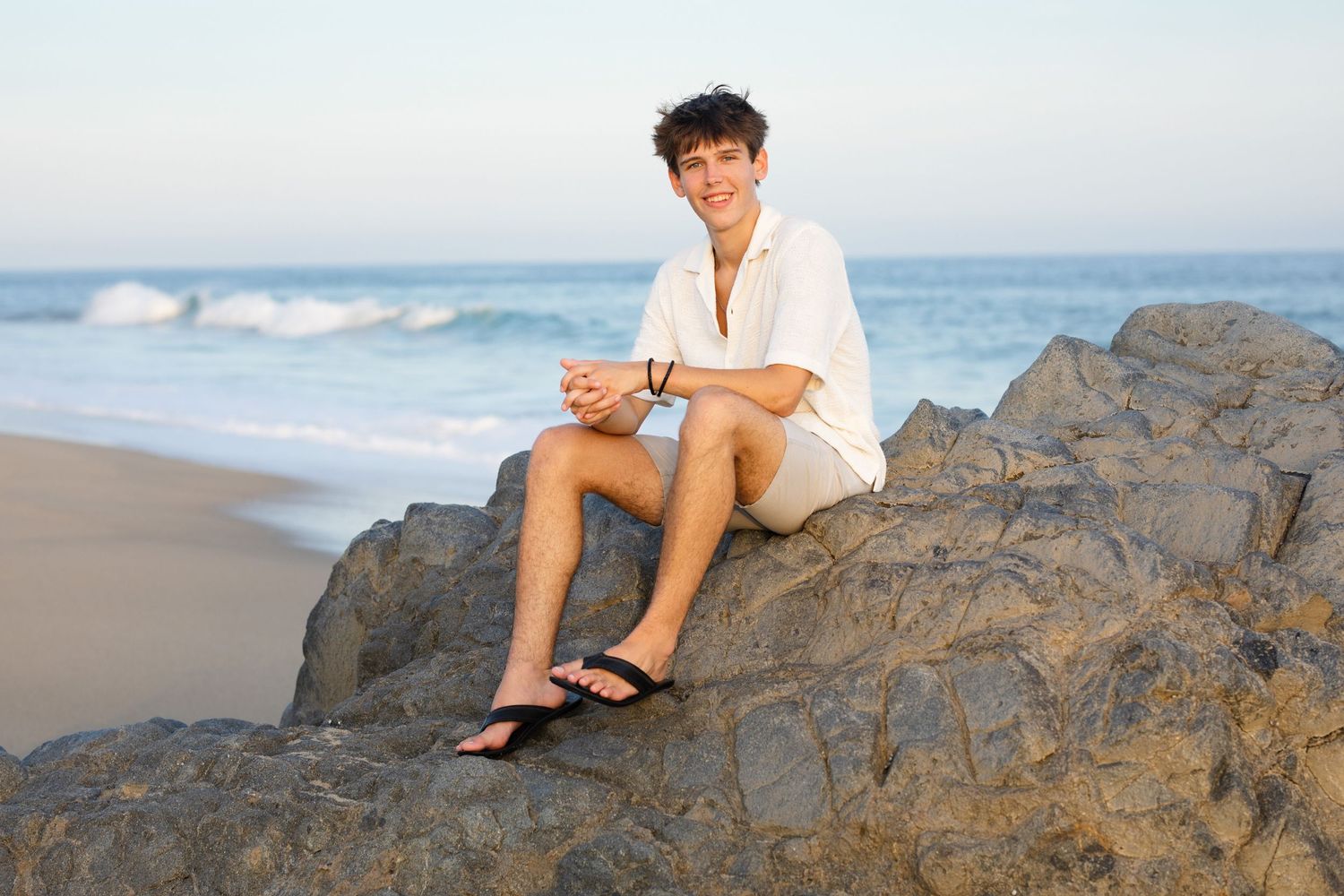 Smiling young man in white shirt sitting relaxed on coastal rocks with ocean waves behind.