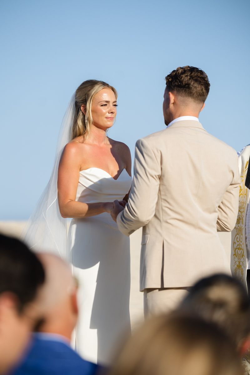 Bride in white strapless gown and veil exchanges vows with groom at beach ceremony.