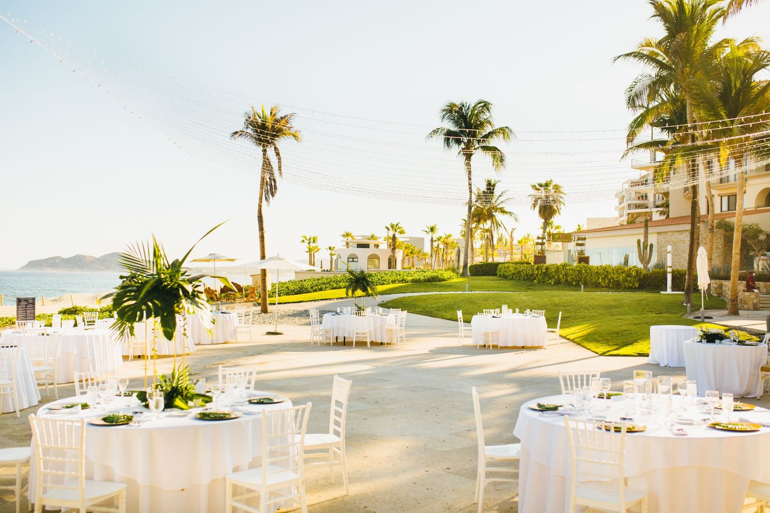 Elegant outdoor wedding reception with white round tables, chiavari chairs, palm trees, and ocean views at a tropical resort.