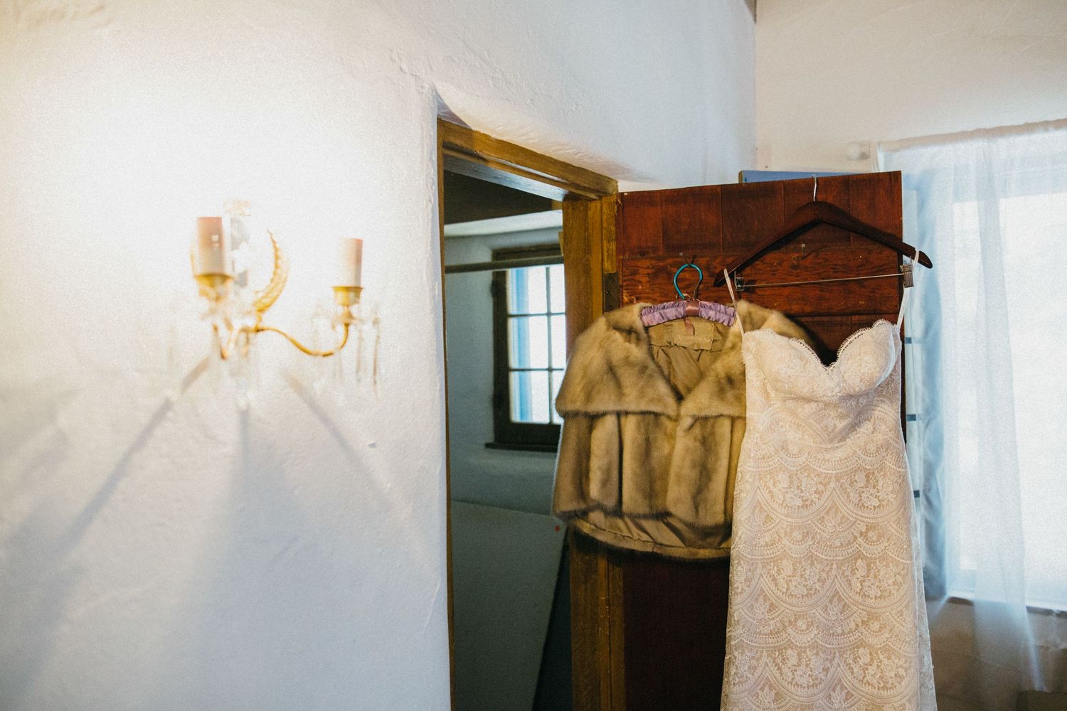 Wedding dress and fur wrap hanging on wooden hangers near a rustic doorway with elegant wall sconce lighting at leo carillo ranch