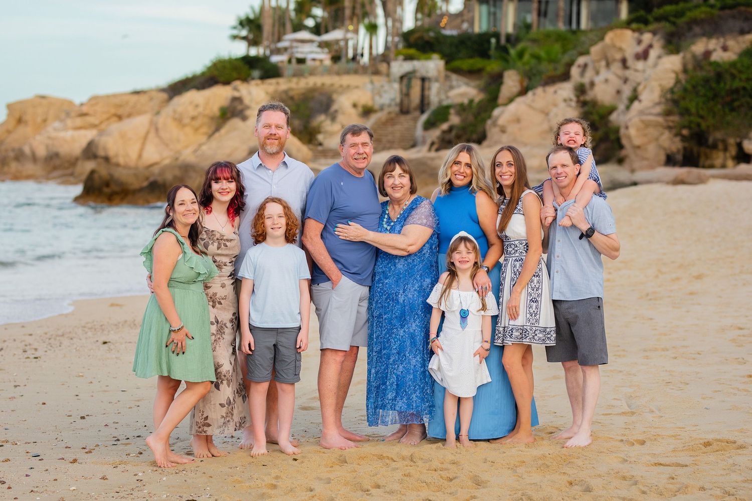 Large family group smiling together on a sandy beach with rocky cliffs in the background.
