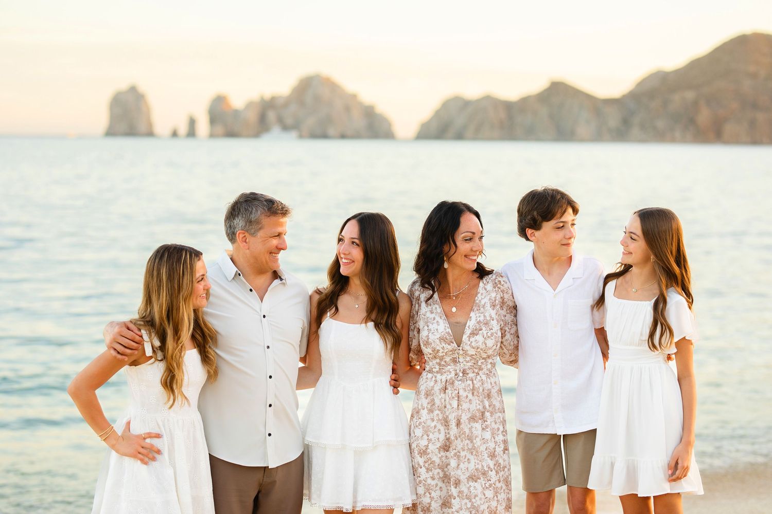 Happy family of six posing together on a beach with rocky cliffs in background.