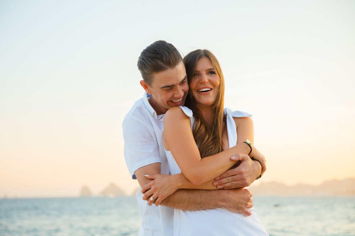Joyful couple embracing and laughing on beach at golden sunset with ocean and rocky cliffs behind.