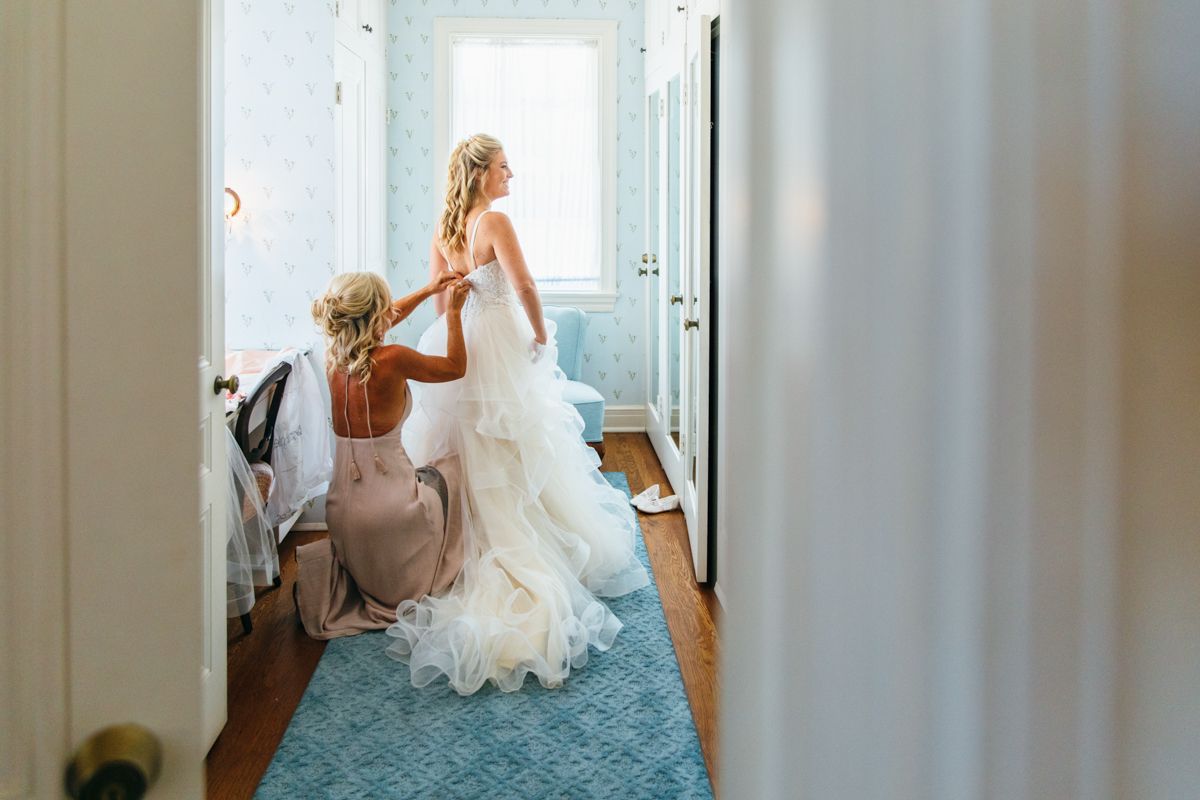 Bridesmaid helps bride button her elegant ruffled wedding gown in a softly lit dressing room.