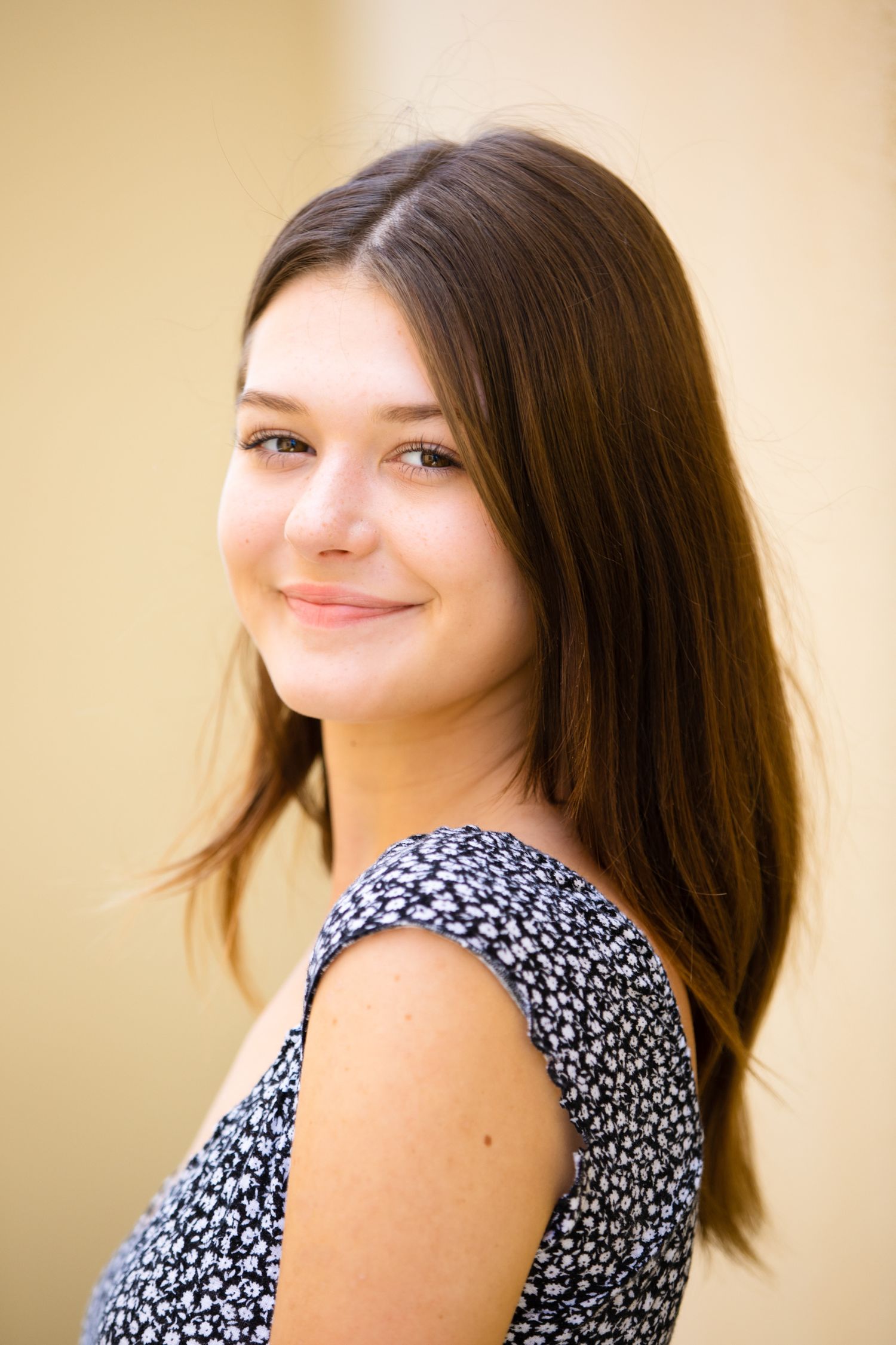 Brunette teenage girl smiling over her shoulder wearing a floral dress against a warm beige background.