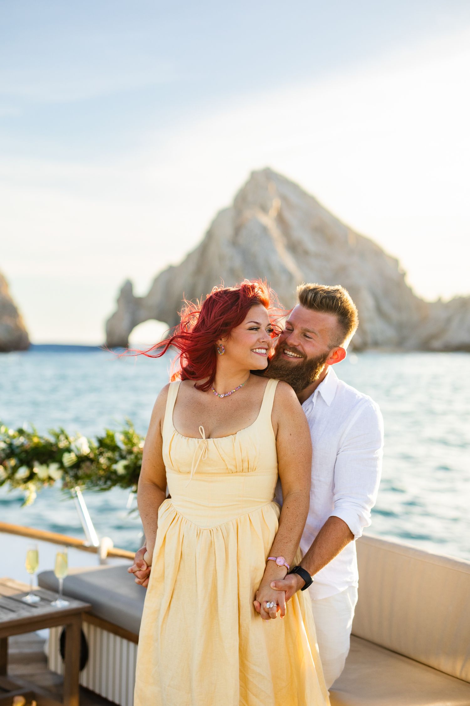 Couple embracing on a boat with red rock arch and ocean backdrop in Cabo San Lucas, woman in yellow dress.