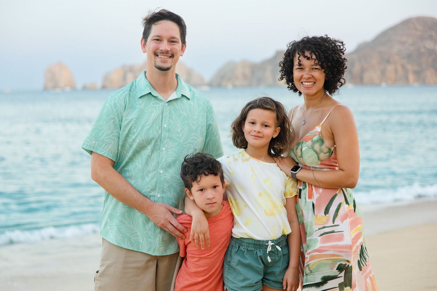 Smiling interracial family of four posing together on a beach with rocky cliffs behind them.