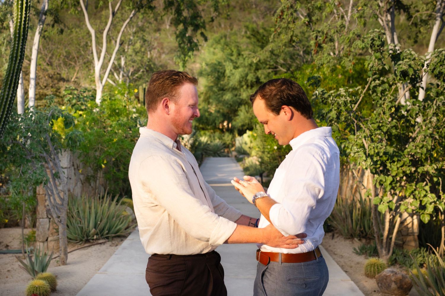 Two men embracing and smiling together on a sunlit garden path with desert plants.