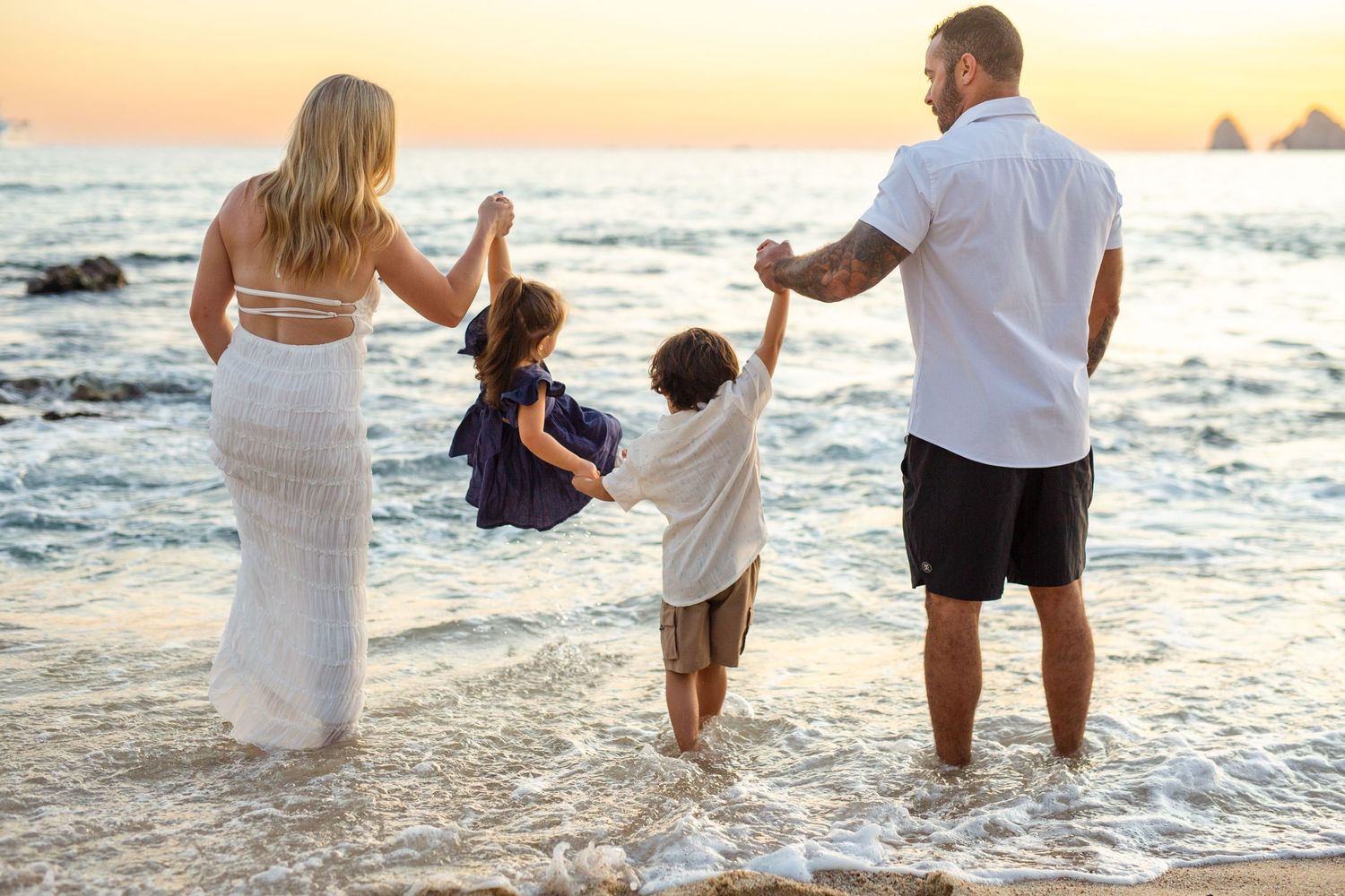 Family of four holding hands at sunset on a beach, parents swinging children over ocean waves in cabo san lucas