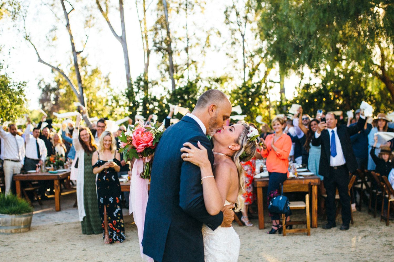 Bride and groom share first kiss at outdoor wedding ceremony surrounded by cheering guests under trees at Leo Carrillo ranch