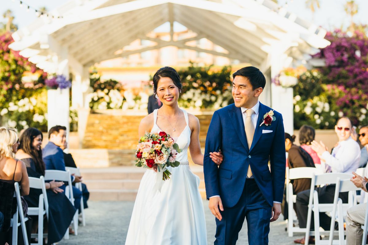 Happy newlyweds walk hand in hand down the aisle at sunny outdoor wedding ceremony.