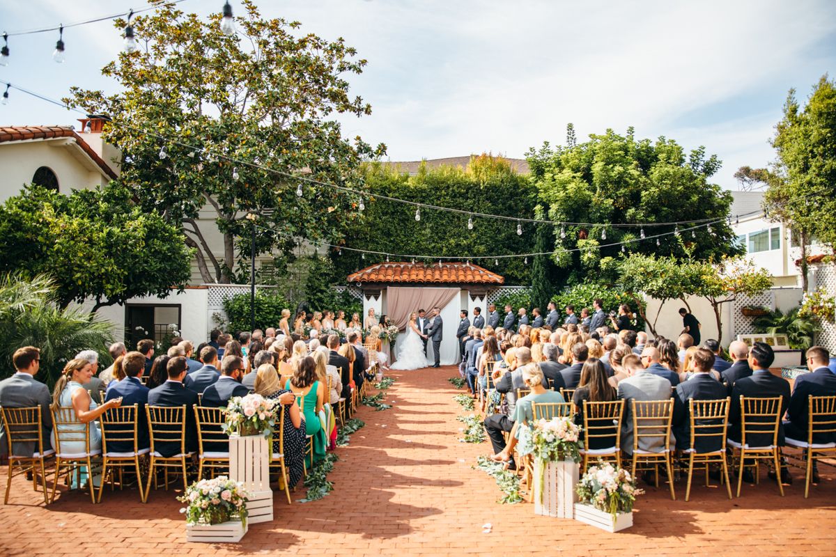 Outdoor wedding ceremony with guests seated in gold chairs on a brick patio surrounded by lush greenery and string lights.