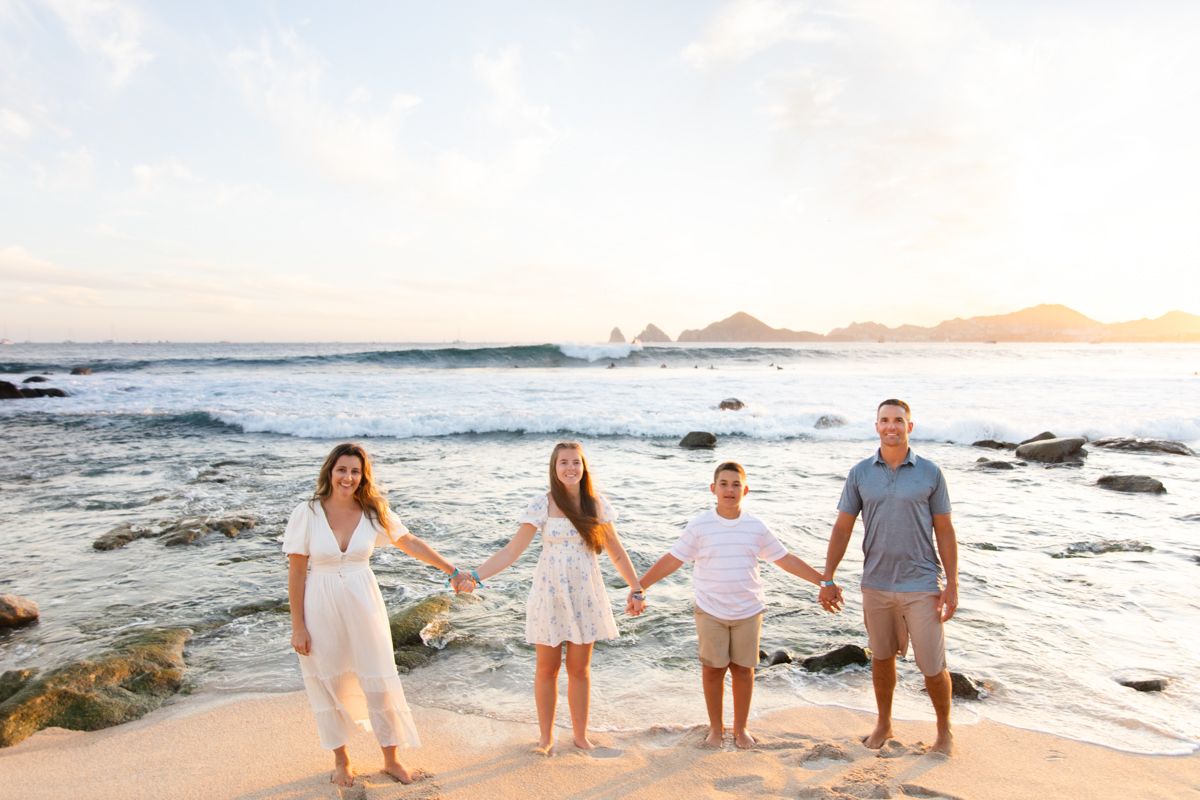 Family of four holding hands in a line on sunny beach with turquoise ocean waves and rock arch behind.