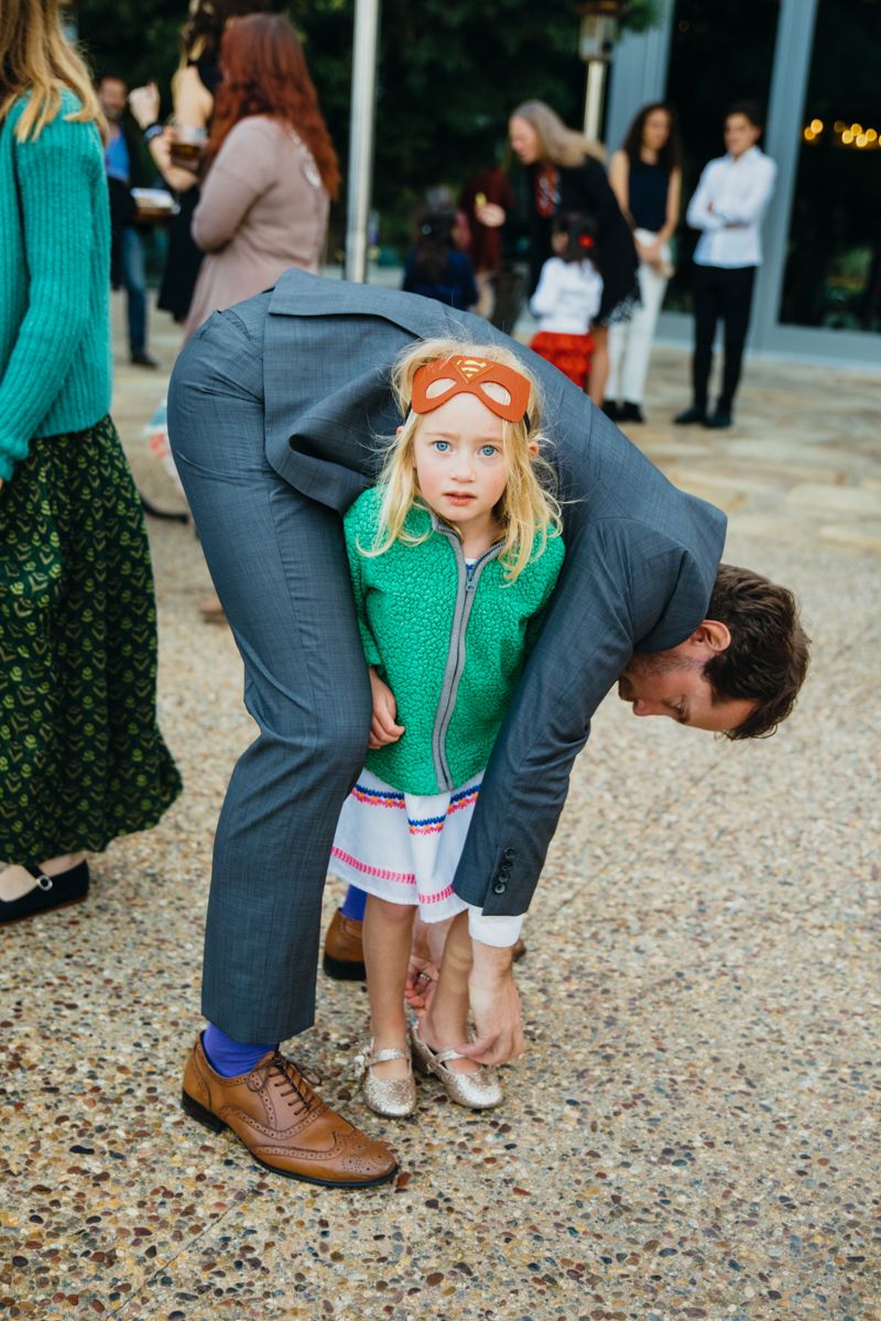 Father ties young flower girl's shoe at outdoor wedding reception venue.