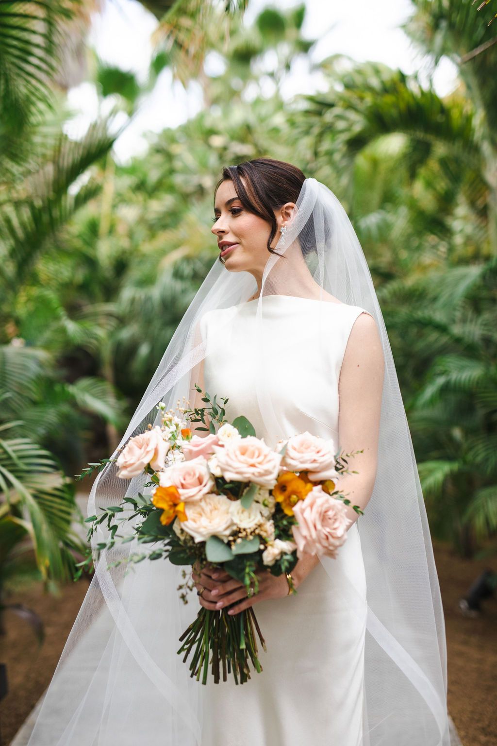 Beautiful bride in white gown with cathedral veil holding pink and orange rose bouquet outdoors.