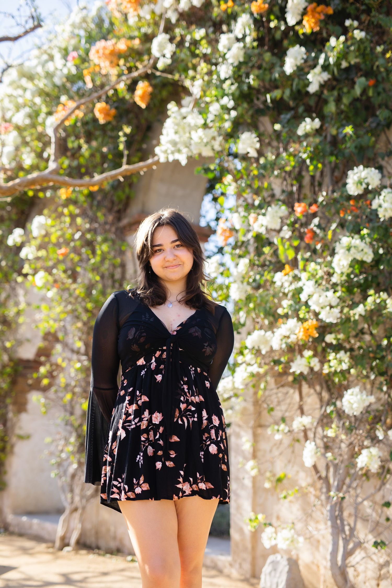 Young woman in black floral romper standing under a blooming flower arch in a sunny garden.