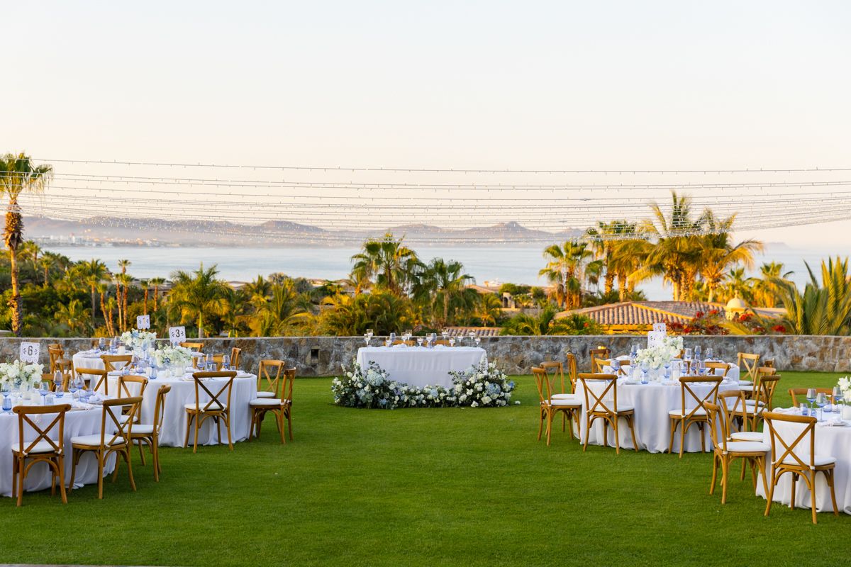 Elegant outdoor wedding reception setup on green lawn with ocean and mountain views at sunset.