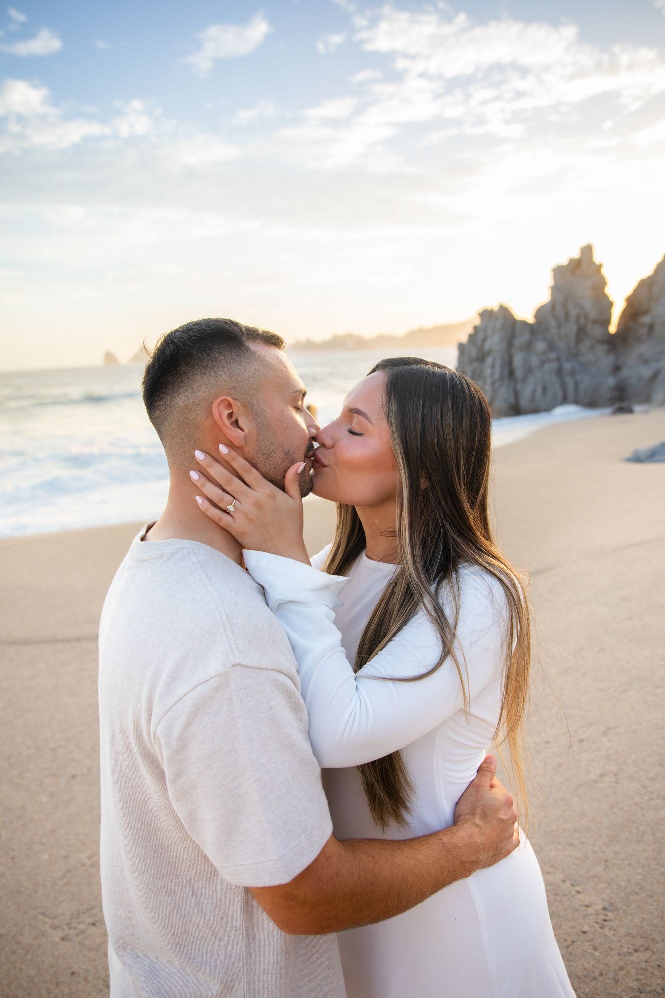 Couple sharing a romantic kiss on a sandy beach at sunset, with rocky cliffs and ocean waves in the background.