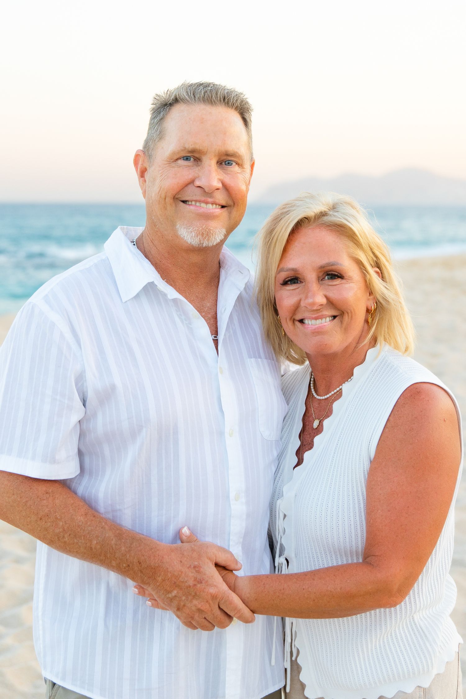 Smiling couple in white outfits posing together on a sunny beach in cabo with ocean and mountains in background.