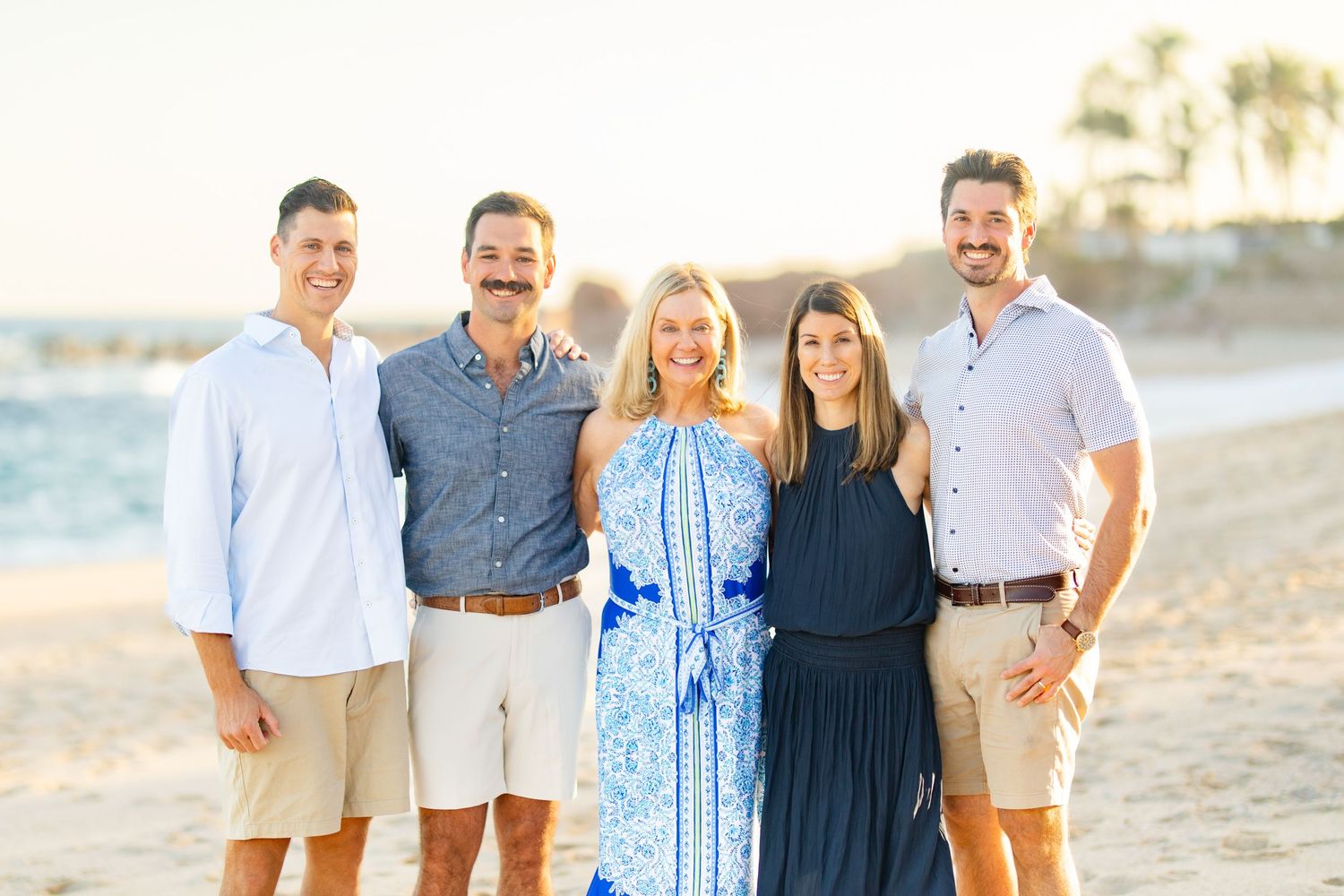 Family of five smiling together on a sunny beach, dressed in casual summer attire with golden hour light.