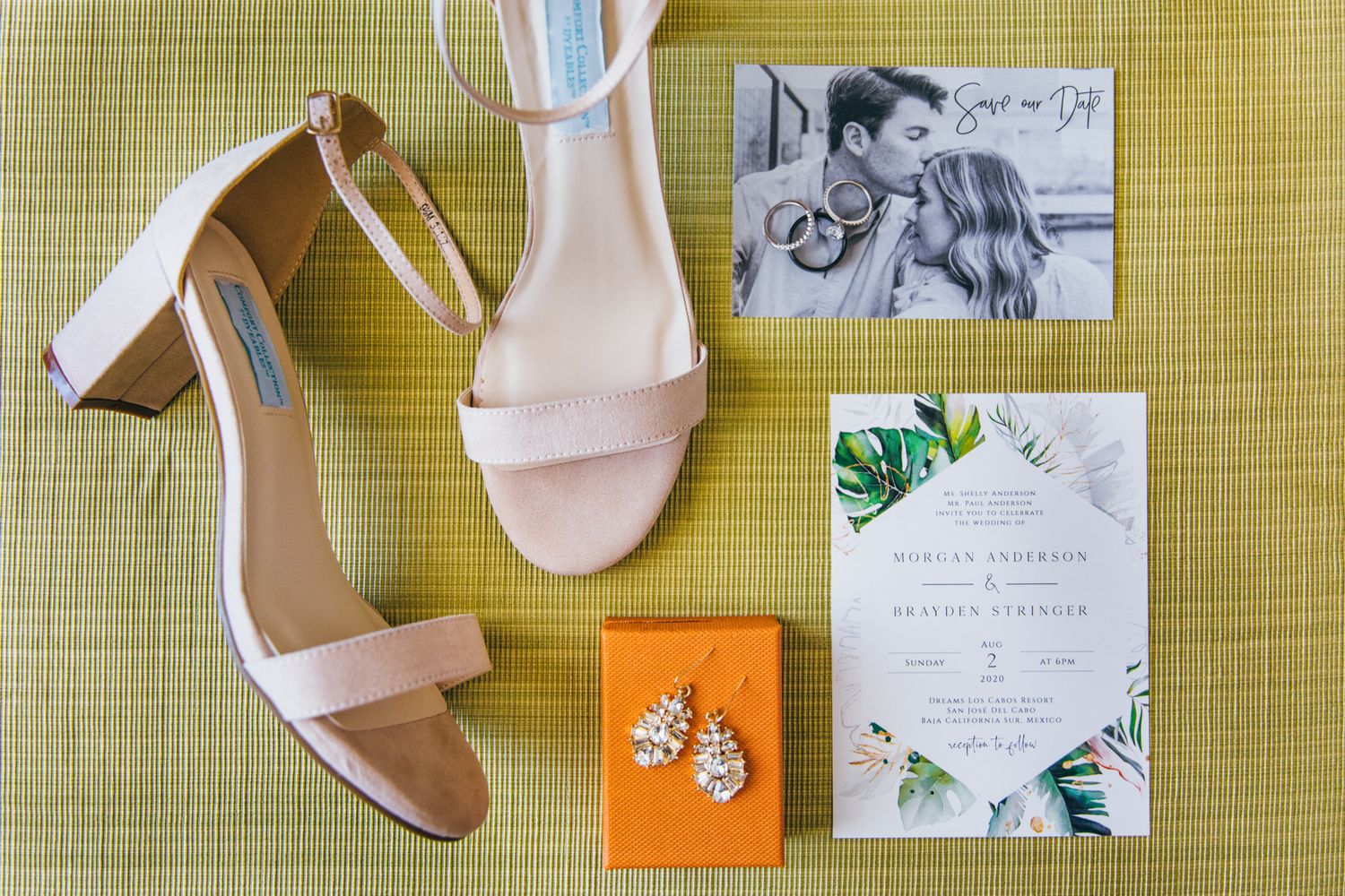Blush pink block heel sandals beside tropical wedding invitation suite and crystal earrings on green textured surface.