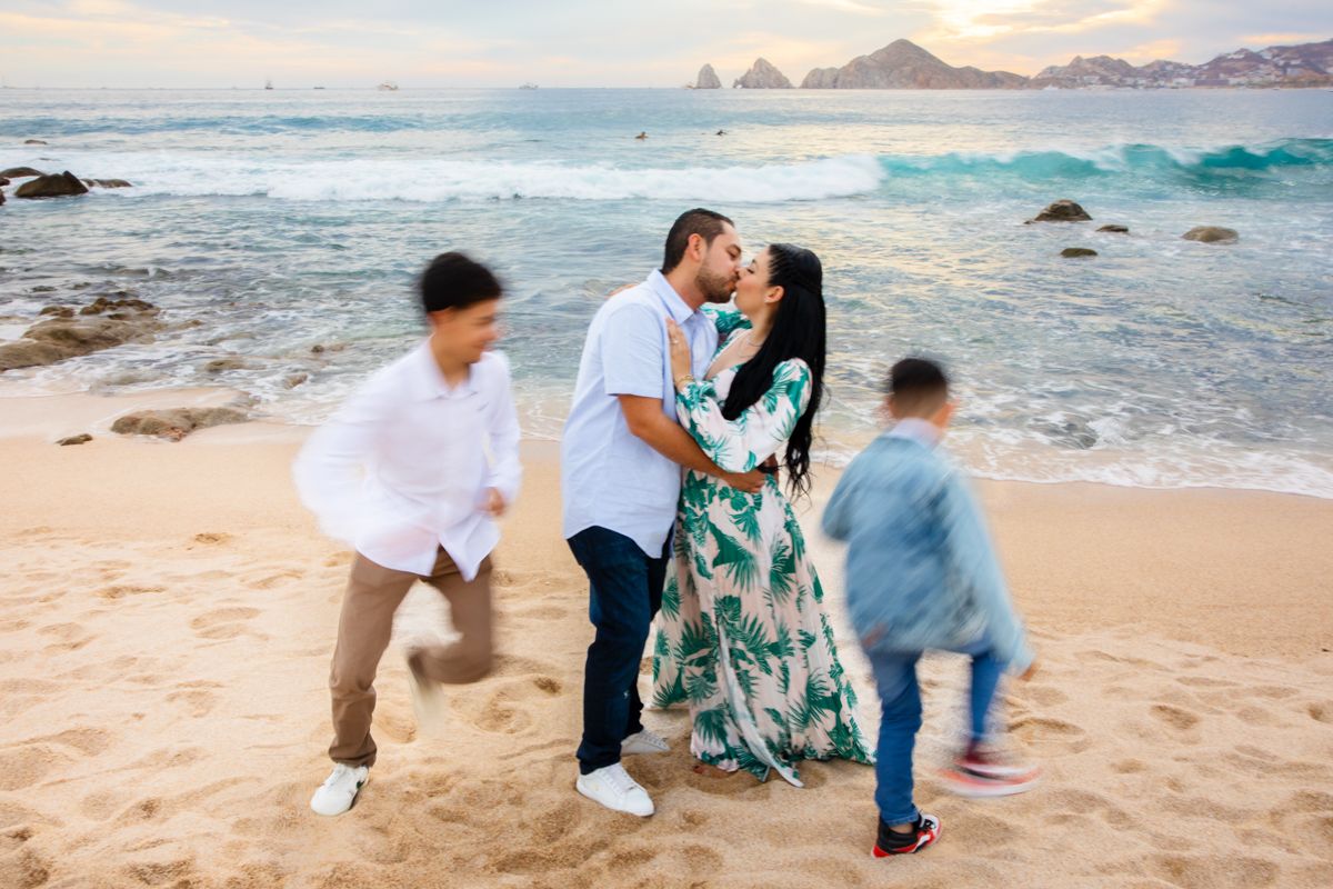 Parents kiss on sandy beach while two children run playfully around them at golden sunset.