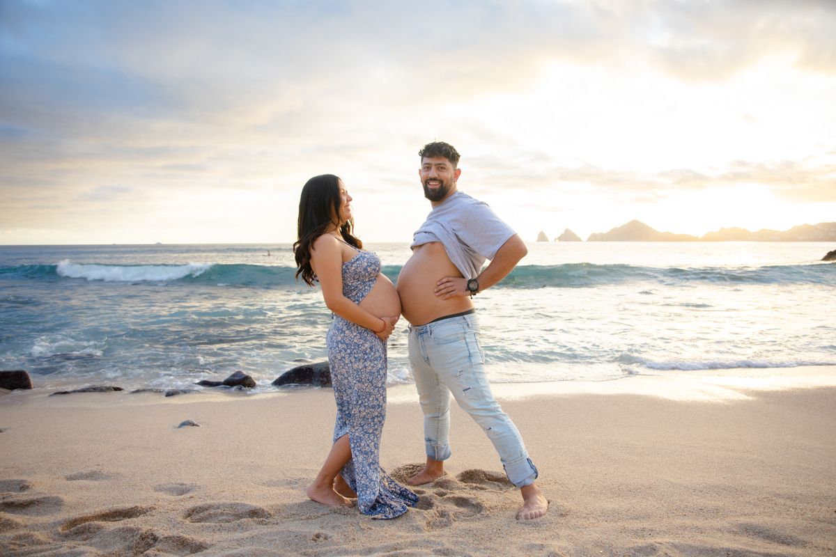 Expecting couple poses on sandy beach at sunset, proudly showing off pregnant belly by the ocean.