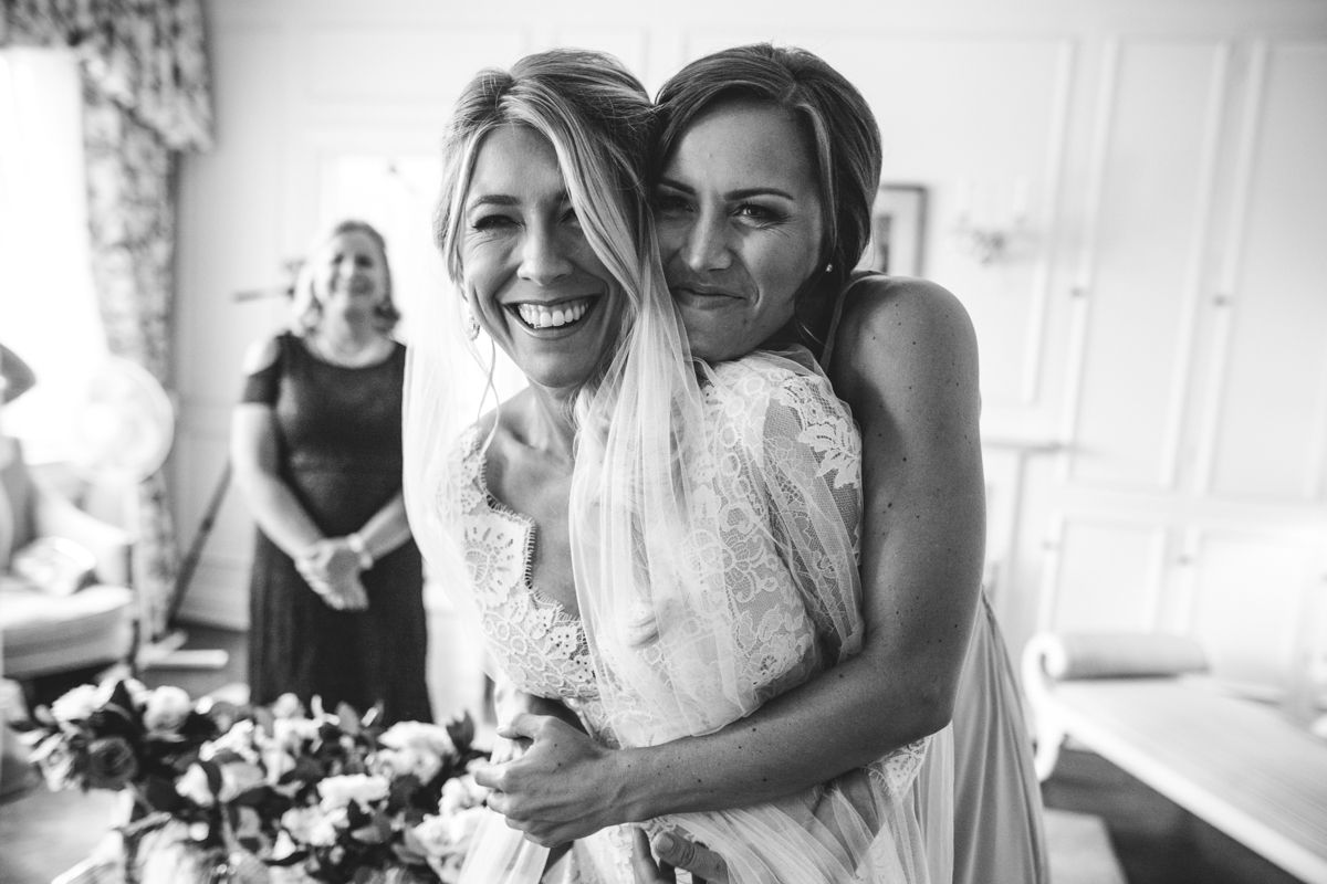 Bride laughing joyfully as bridesmaid hugs her tightly in black and white wedding photo.