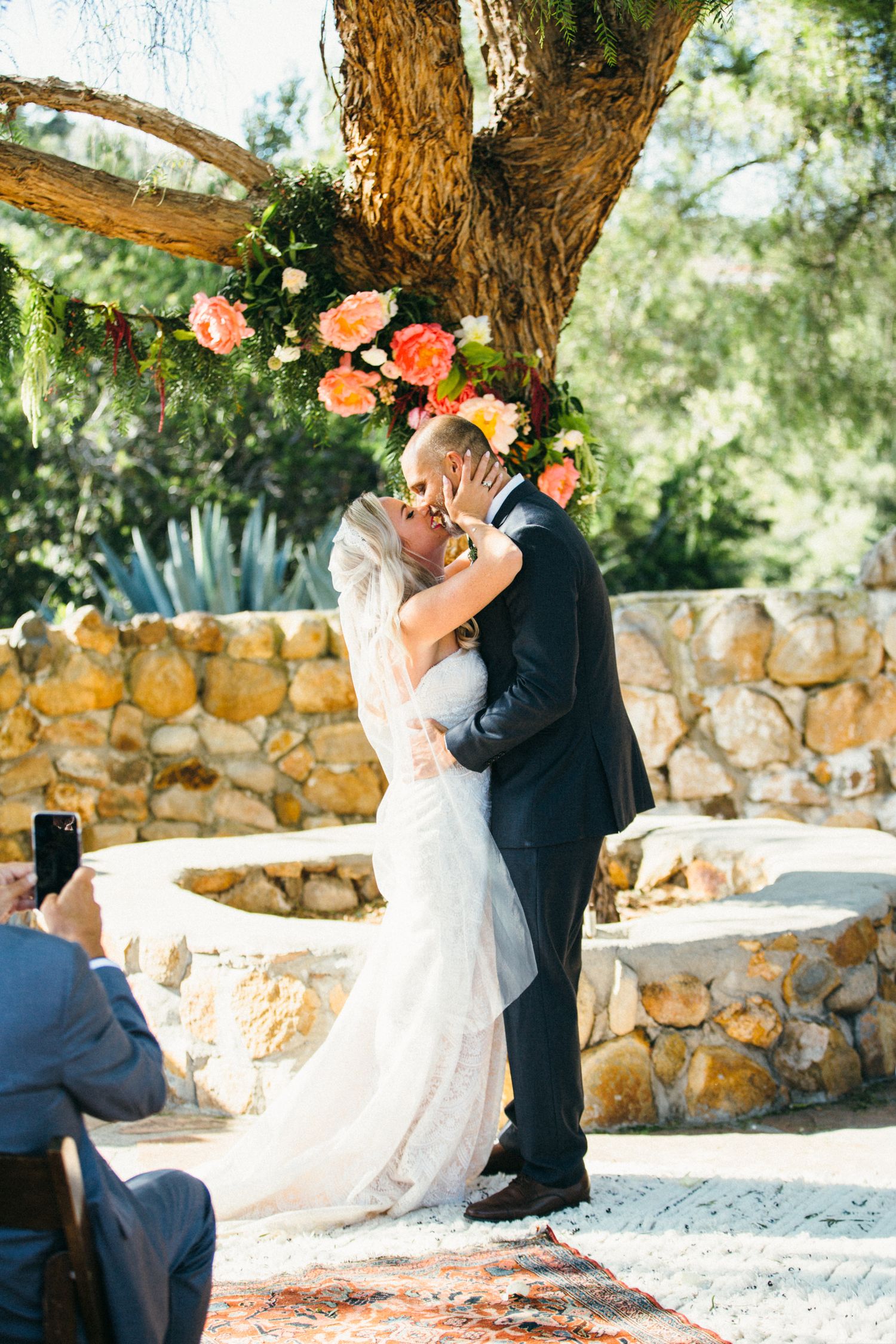 Bride and groom share first kiss at outdoor wedding ceremony under floral arch with a stone wall backdrop at Leo Carrillo ranch