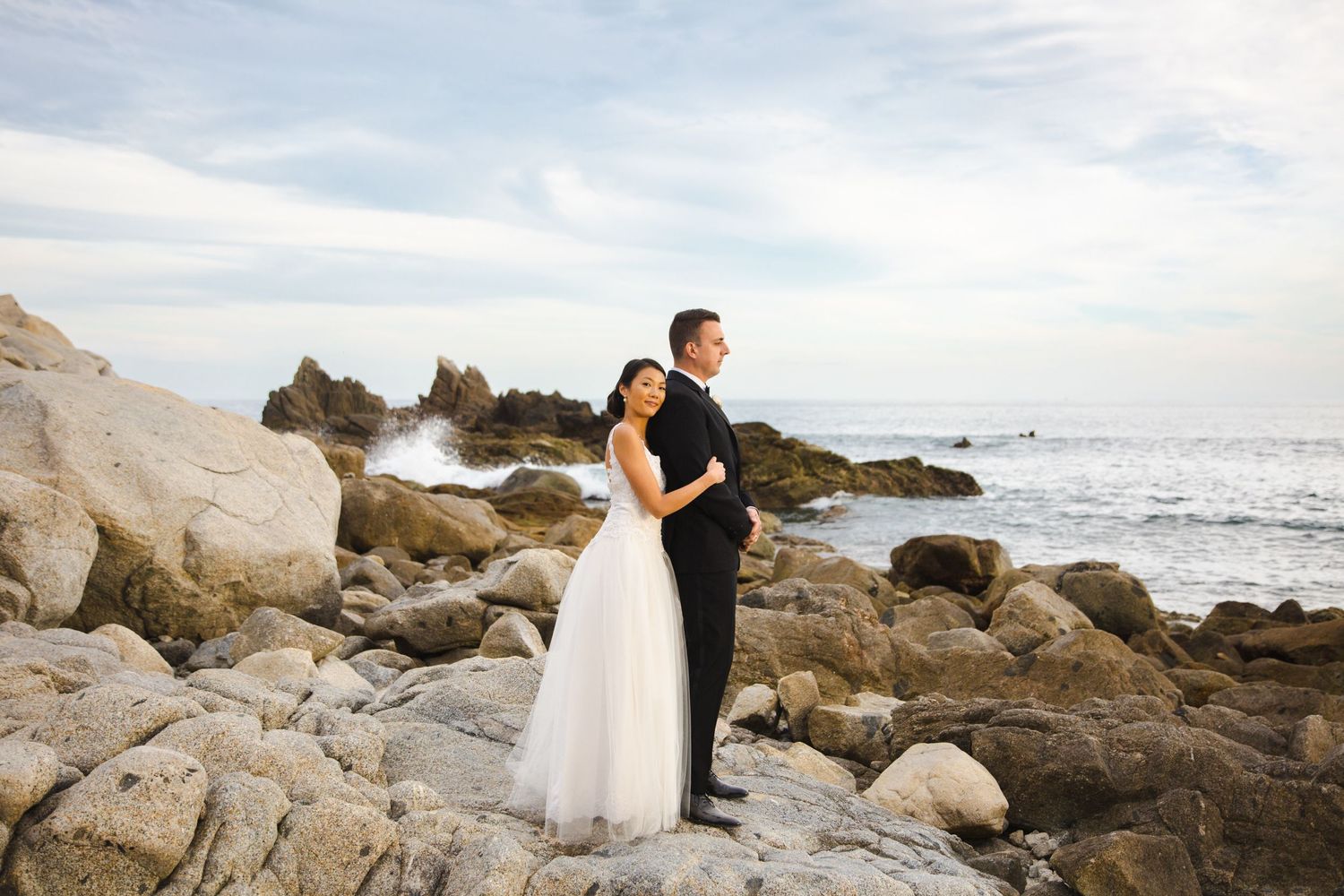 Bride and groom stand back-to-back on rocky coastal shore, waves crashing behind them under a cloudy sky.