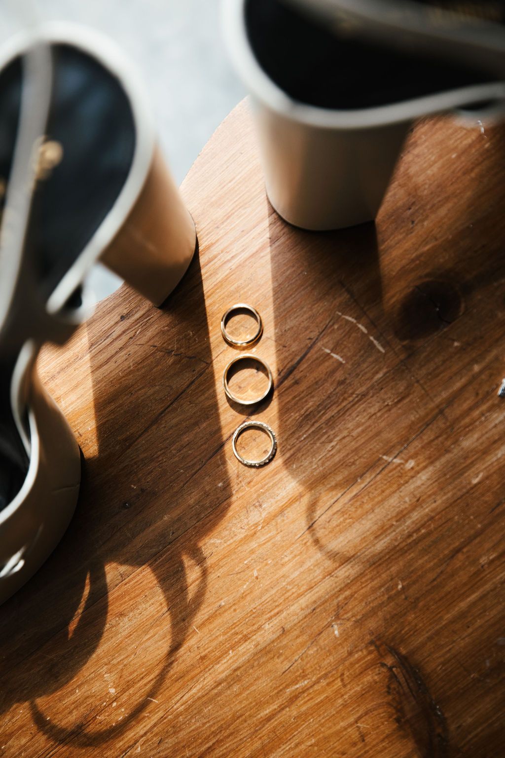 Three gold wedding bands arranged in a row on a wooden surface beside white bridal shoes.