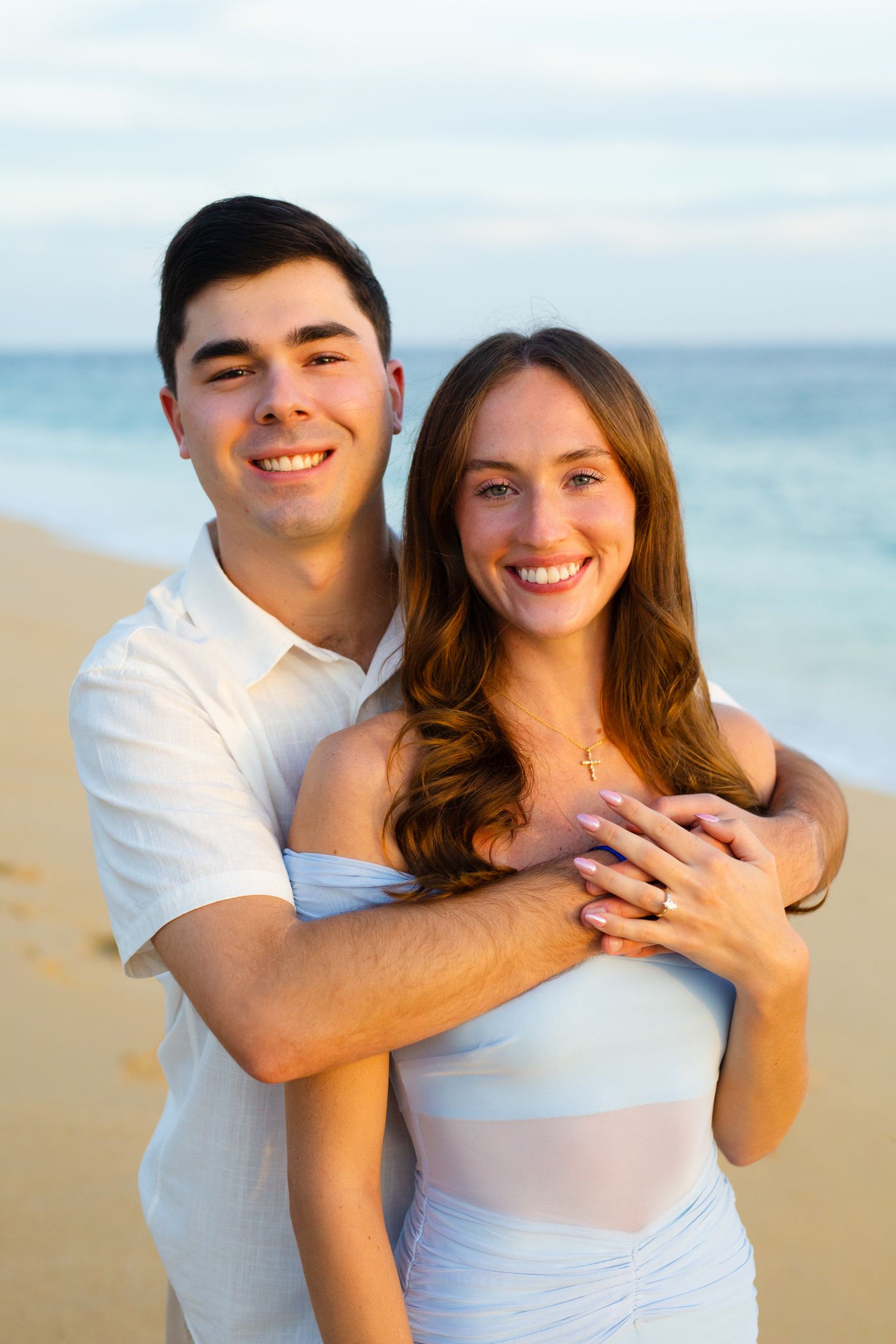 Happy couple embracing on a sandy beach at sunset, wearing white outfits with ocean waves in the background.
