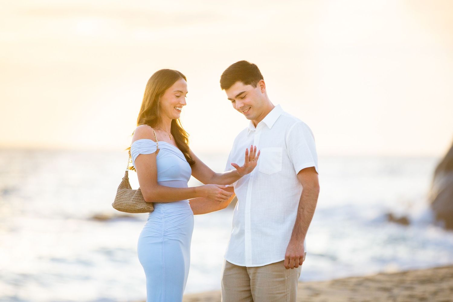 Couple sharing a tender moment on a beach in cabo at sunset, woman in blue dress admiring engagement ring.
