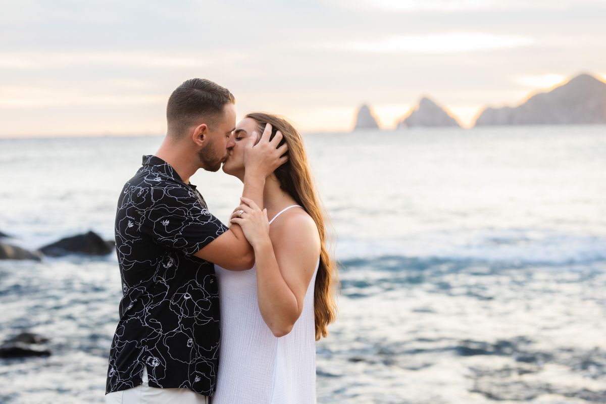 Couple shares a tender kiss on a rocky beach at sunset with ocean waves and rock formations behind.