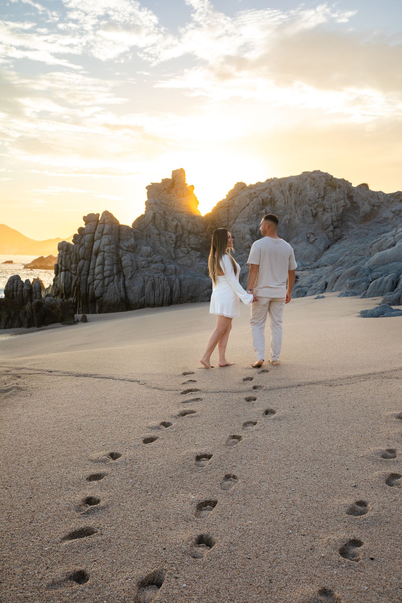 Couple holding hands walking on sandy beach at sunset with rocky formations and footprints trailing behind them.