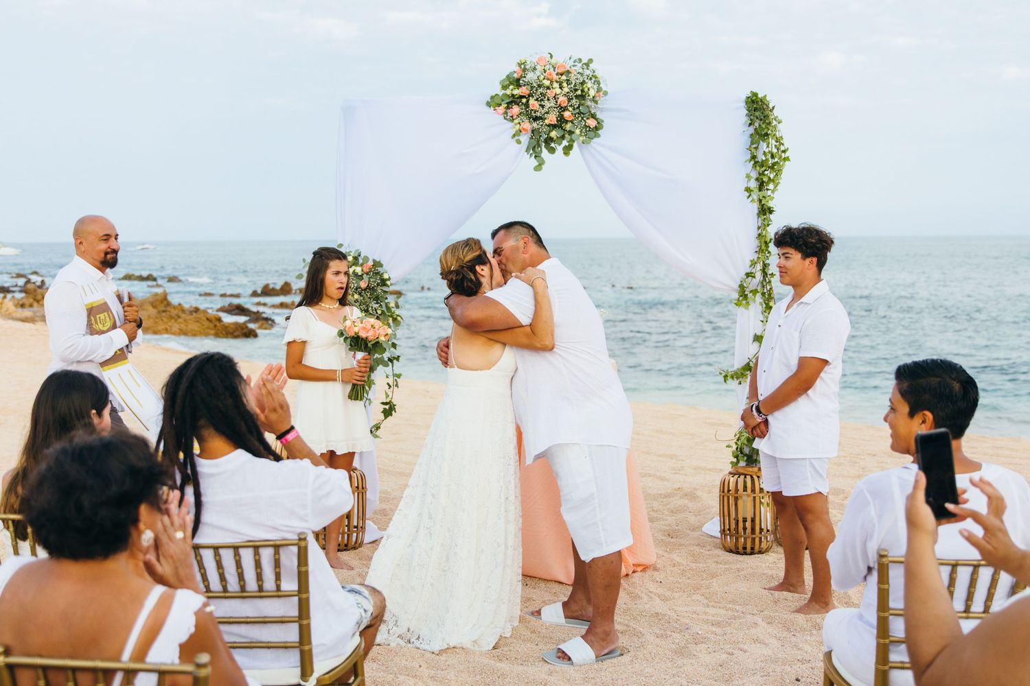 Couple shares first kiss at romantic cabo beach wedding ceremony with white decor, floral arch, and guests seated on golden chairs.