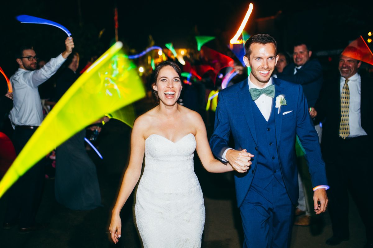 Joyful newlyweds exit reception through crowd of guests waving colorful glow sticks at night.