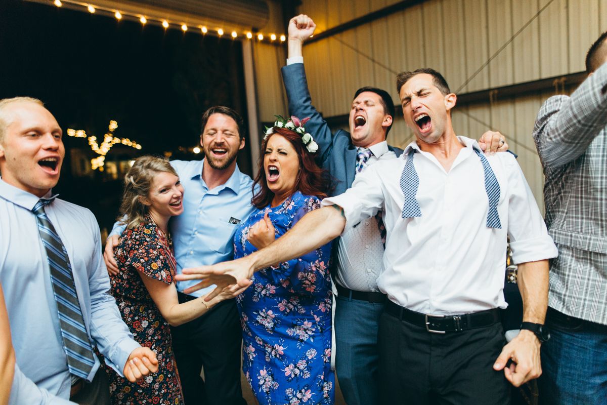 Excited wedding guests cheer and celebrate together in a barn venue with string lights overhead.