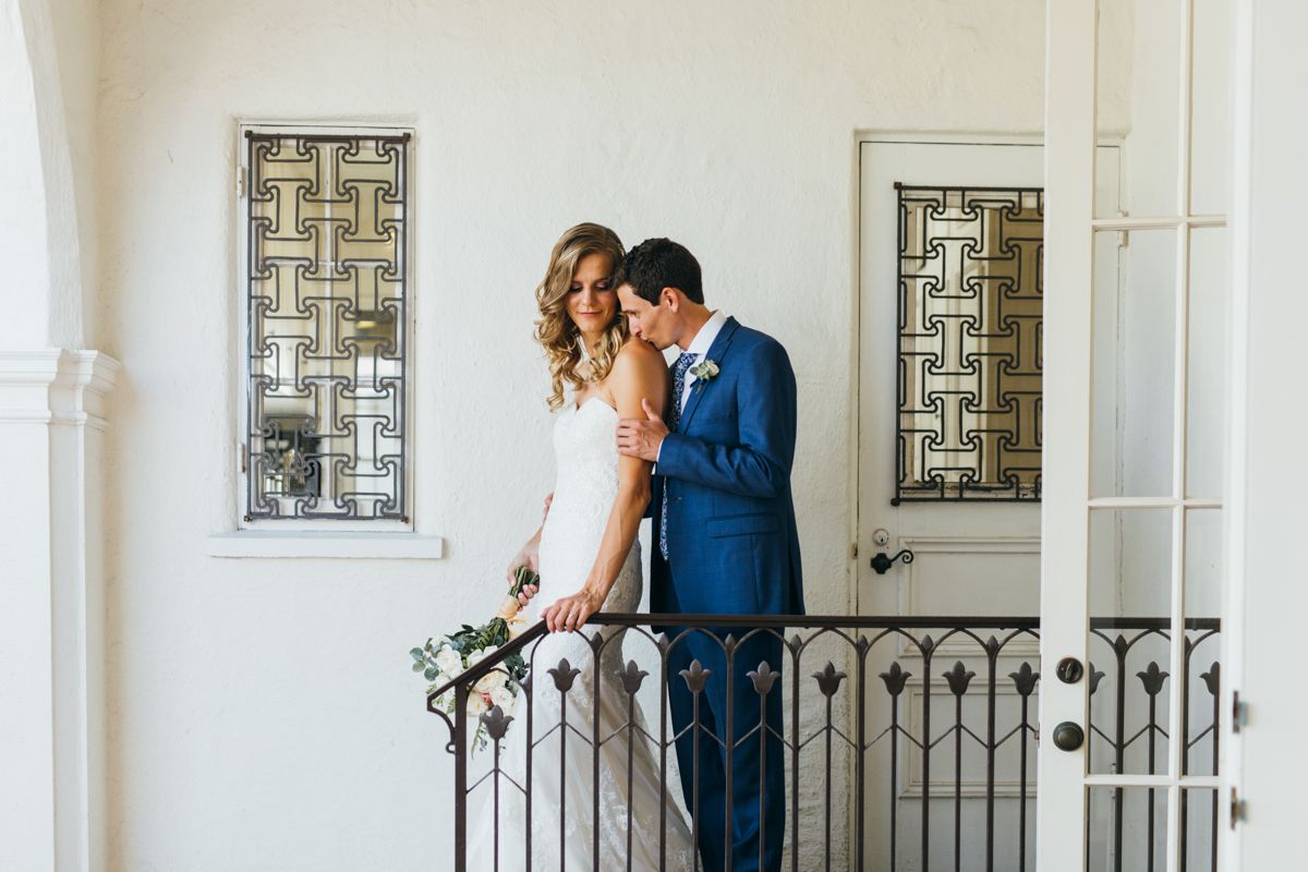 Bride and groom share a romantic moment on an ornate iron balcony with decorative windows.