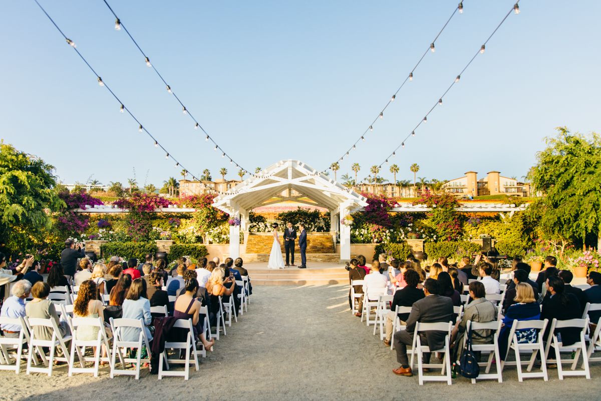 Guests seated in white chairs watch wedding ceremony at elegant outdoor venue with pergola and string lights.