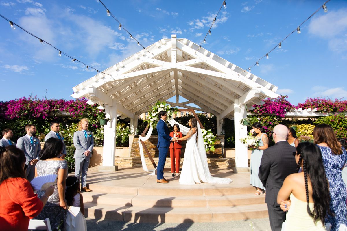 Outdoor wedding ceremony under white pergola with string lights, colorful bougainvillea, bride and groom exchanging vows.