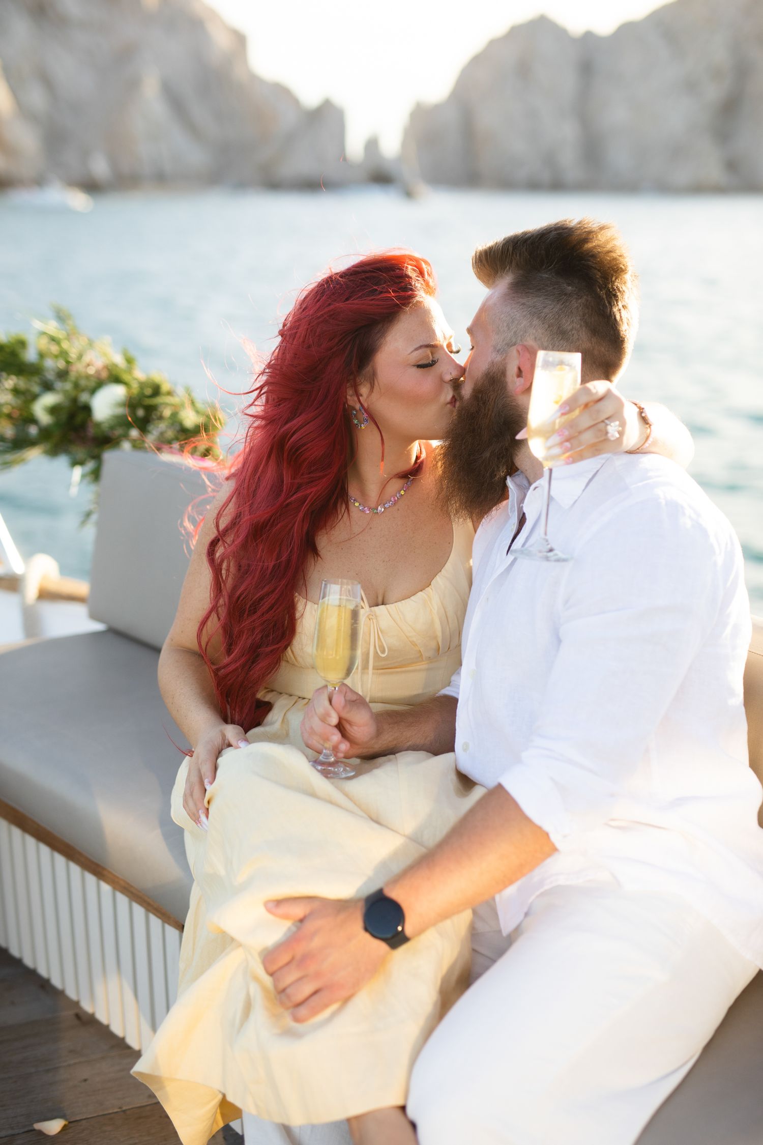 Couple kissing on a boat in Cabo San Lucas, woman with red hair in yellow dress, man in white holding champagne glasses.