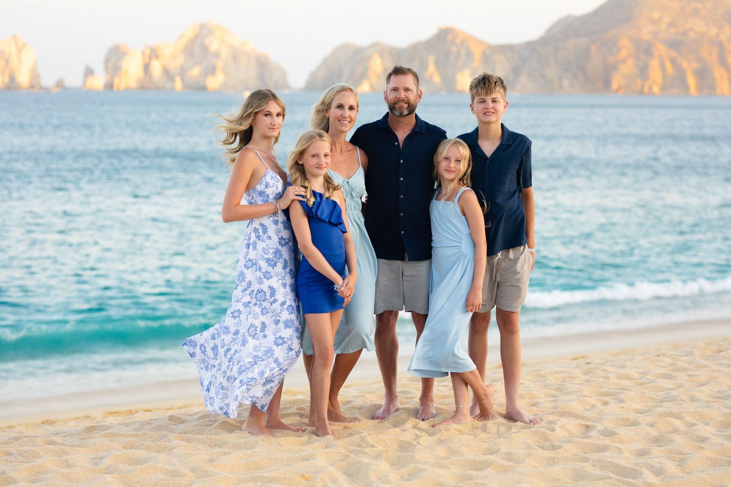 Family of five posing together on a sandy beach in Cabo San Lucas with rocky cliffs and turquoise ocean in the background.