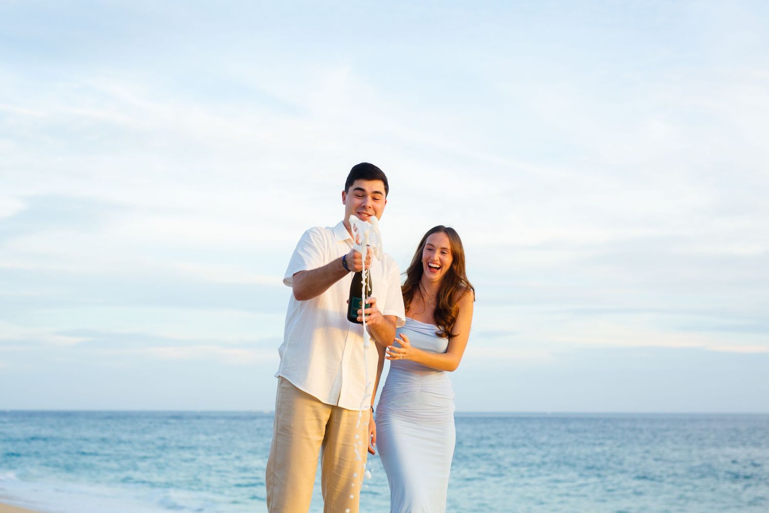 Joyful couple celebrating beach proposal with champagne bottle by the ocean under cloudy sky.