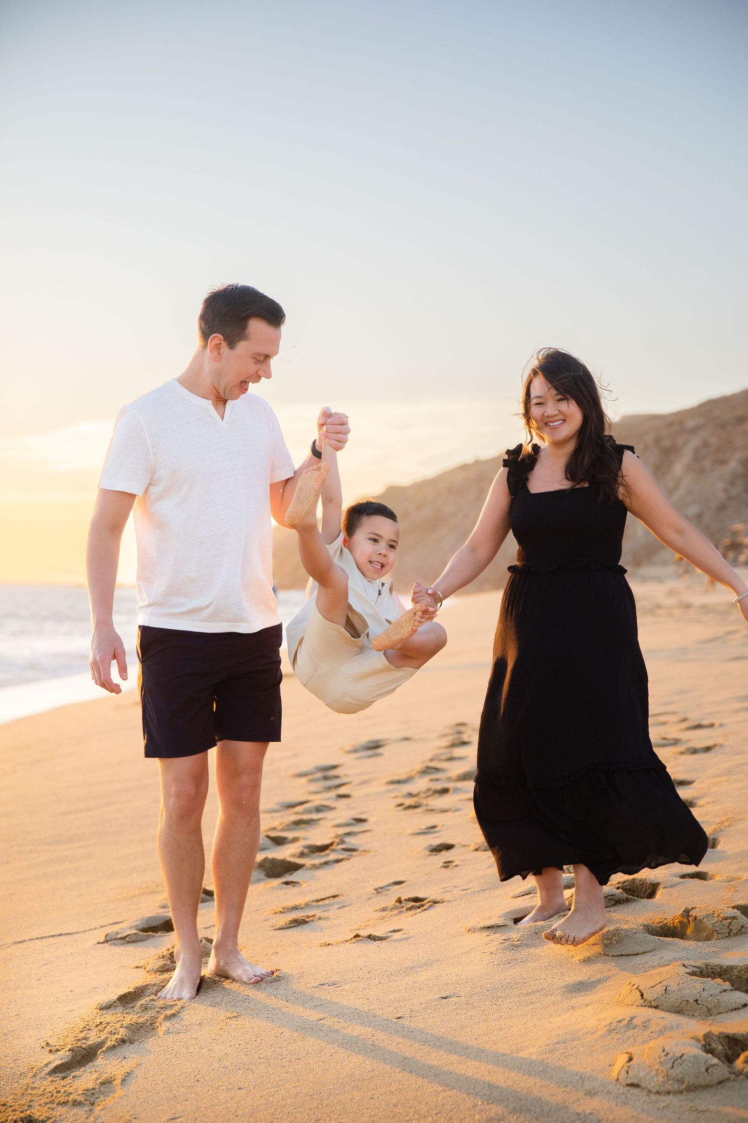 Family of three enjoying a sunset beach walk in Cabo San Lucas, parents swinging their child between them on golden sand.