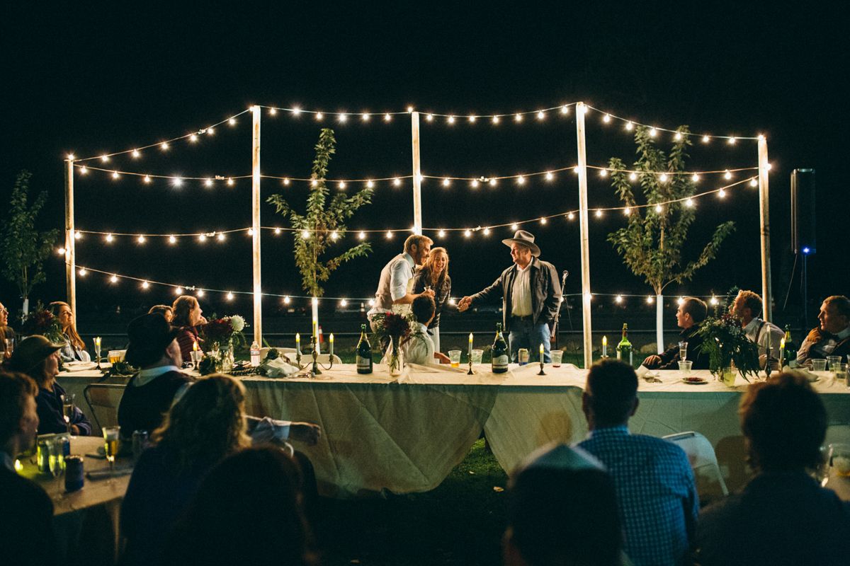 Couple cuts wedding cake at elegant outdoor reception under string lights at night with guests.