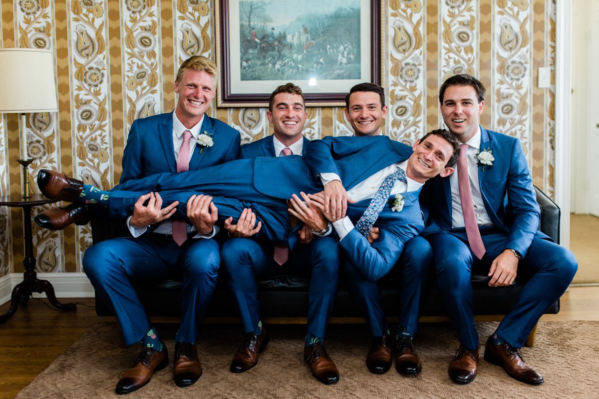 Four groomsmen in blue suits playfully pose on a sofa, holding one groomsman horizontally in a fun photo.